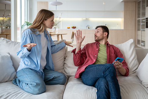 Aggressive wife with husband trying to talk gesturing hands. Family couple having misunderstanding showdown scandal sitting on couch at home. Neurotic relationships, toxic relations, marital discord.