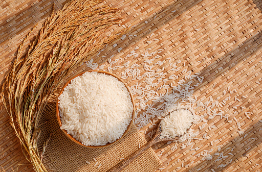White rice (Thai Jasmine rice) in ceramic bowl and wooden spoon with ear of paddy on threshing basket background, top view with copy space