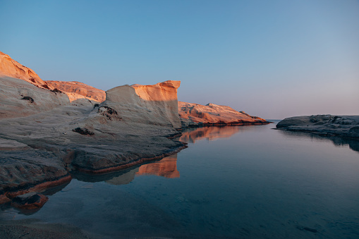 A serene moment as dawn breaks over the stunning breathtaking white volcanic cliffs beauty of Sarakiniko Beach on Milos Island as the first light of dawn illuminates the rugged coastline. The golden hues of sunrise cast a warm glow over the unique white volcanic cliffs and turquoise waters of the Cyclades Sea, creating a scene of unparalleled natural beauty in Milos, Greek Islands, Greece