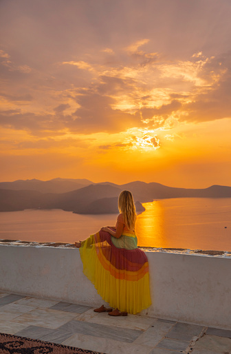 A young tourist woman sits in contemplation against the backdrop of the rising sun, horizon, and ocean at the enchanting Panagia Korfiatissa Church in Plaka village, Milos Island, Cyclades, Greece. As the sun ascends, casting its warm glow over the tranquil seascape, she finds solace and wonder in the serene beauty of the moment. This idyllic scene captures the essence of peaceful reflection amidst the natural splendor of Milos Island, inviting viewers to pause and embrace the beauty of the sunrise.
