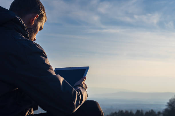 istock Young man in jacket using tablet. Landscape with distant hill at sunrise, copy space sky 2076025658