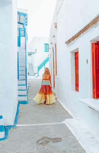 Back view of a young beautiful happy traveller female lady with a red dress and bag enjoys the scenery and walking through the traditional white and blue small, whitewashed alleys of Mikonos town, Cyclades islands of Greece during summertime.