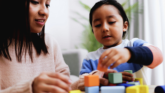 Latin American mother and son child having fun playing games with wood toy bricks at home. Education and family leisure time concept