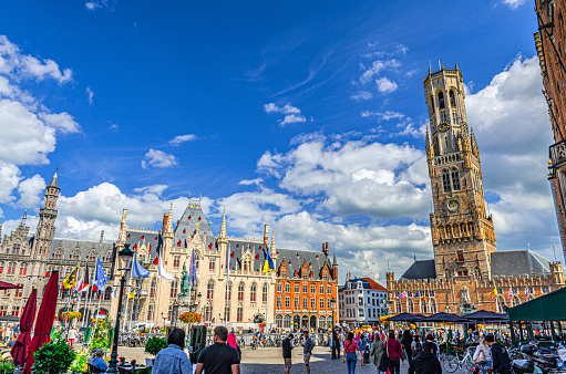 Bruges, Belgium, July 5, 2023: crowd of many people tourists walking down Markt Market square, Belfry of Bruges Belfort bell tower and Provincial Court in Brugge old town historical city centre