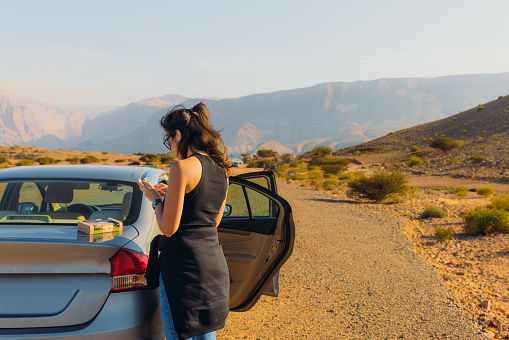 Side View of Middle Eastern female in eyeglasses enjoying the lunch break by her rental car during journey in the mountains of Oman during sunny day