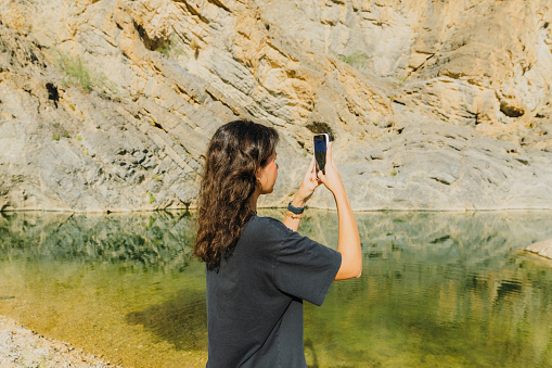 Side view of arabian female with black hair wearing smart watch making photos of the river hidden In the picturesque canyon in Oman