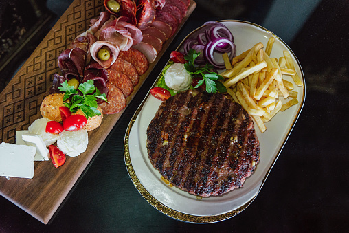 Grilled balkan meat pljeskavica with french fries and traditional salami, cheese and specialties on a black table