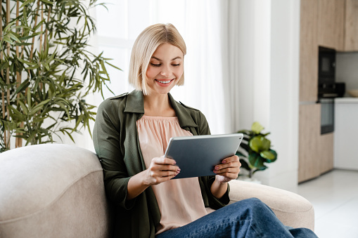 Smiling caucasian freelancer using digital tablet for online shopping from home. Female woman girl watching video movie relaxing on sofa couch