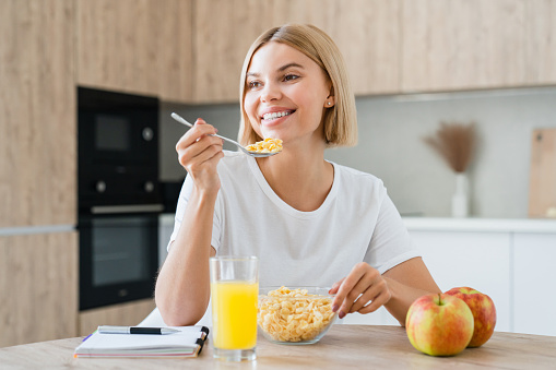 Smiling young woman full of energy for breakfast. Girl eating cereals drinking orange juice with apples at home kitchen in the morning. Healthy eating habits concept