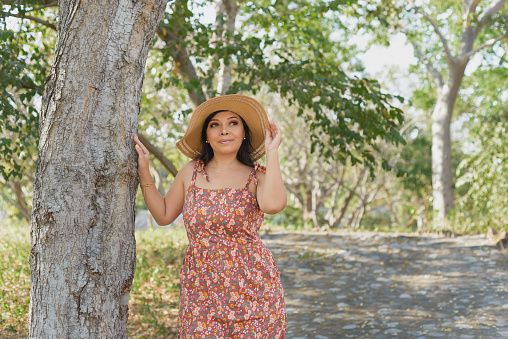 Woman wearing hat and dress with flowers admiring nature in a public park close to a tree. Sunny summer day.