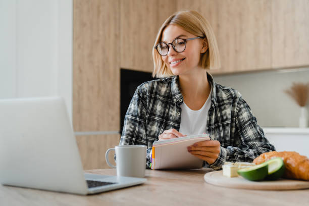 Happy young woman taking notes while working on laptop at home kitchen.