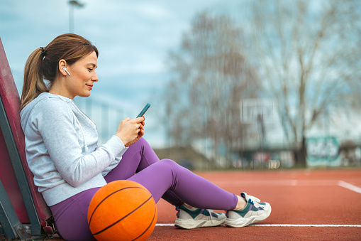 Seated on the ground of the basketball court, the woman finds solace in her mobile phone, enjoying a peaceful break from her activities as she takes a moment to unwind