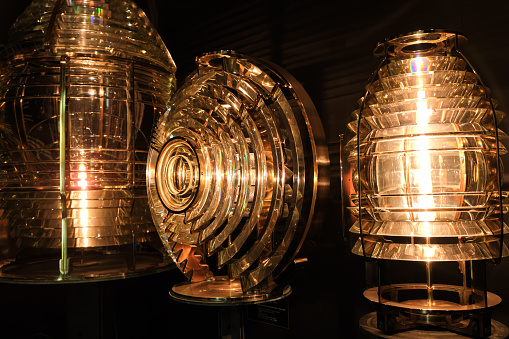 Close image of the glass prisms making up Fresnel lens with lights on. Used in lighthouse beacons and lanterns to concentrate light from compact lamp into revolving light beams or flashes. Three different shapes, different orders, dark room.