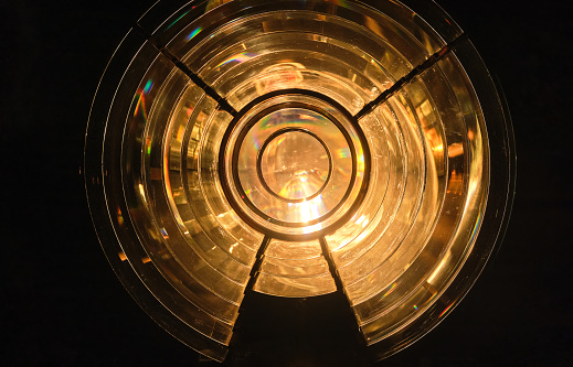 Close image of the glass prism making up Fresnel lens with lights on. Used in lighthouse beacons and lanterns to concentrate light from compact lamp into revolving light beams or flashes. Round shape, dark room, lights on.