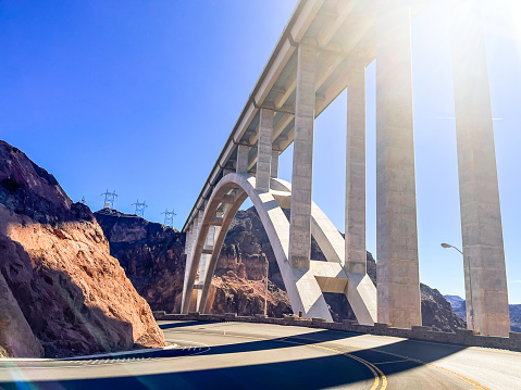View of the arched Hoover Dam Bridge at early morning from the roadway below with sunlight, shadow, blue sky, red rocks.