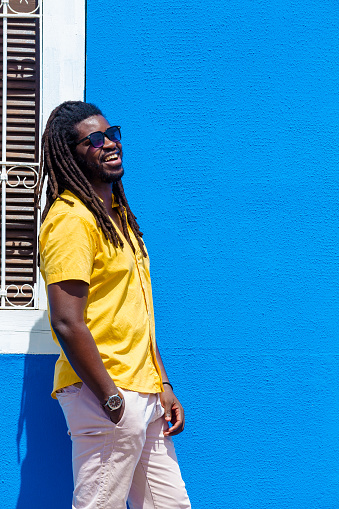 black man wears a yellow shirt, sunglasses, white pants, smiles and has his hands in his pockets in front of the facade of an old blue house