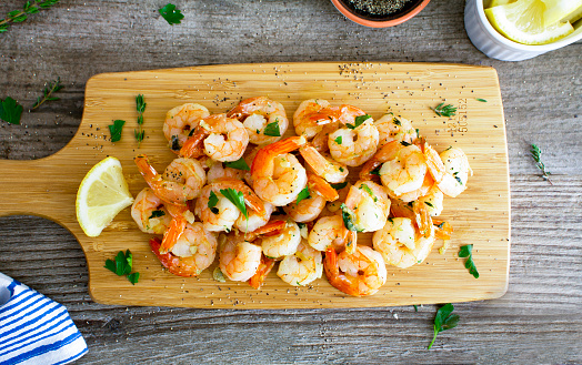 Fried shrimp with garlic and herbs served on a wooden chopping board with lemon and herbs.\nShot from above on a rustic wooden table.