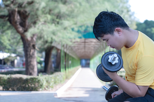 Asian chubby teenboy doing exercise with dumbbells in outdoor park in late afternoon of the day.