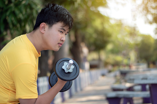 Asian chubby teenboy doing exercise with dumbbells in outdoor park in late afternoon of the day.