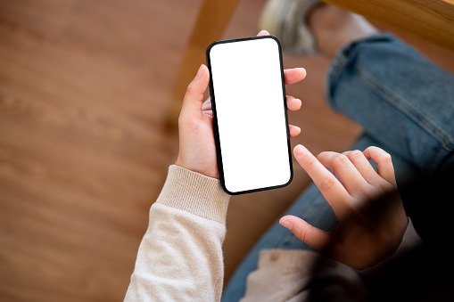 A close-up image of a woman using her smartphone at a table in a cozy cafe, featuring a white screen smartphone mockup to display your graphic ads on the screen. people and wireless technology