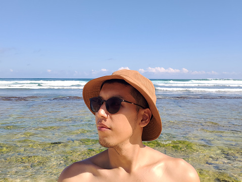 Young Asian man wearing hat and black glasses on the sunny summer beach trip