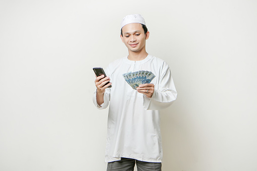 happy muslim man holding money rupiah banknotes and  mobile phone. People religious Islam lifestyle concept. celebration Ramadan and ied Mubarak. on isolated background
