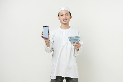happy muslim man holding money rupiah banknotes and showing mobile phone blank screen. People religious Islam lifestyle concept. celebration Ramadan and ied Mubarak. on isolated background