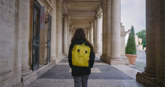 A lone woman with a backpack walking at the entrance of a grand historical building framed by classical columns, evoking a sense of exploration and history.