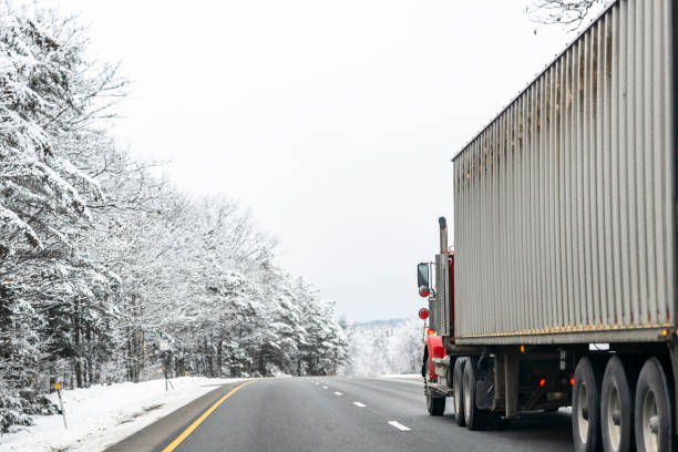 istock Red classic day cab big rig semi truck transporting cargo in long container driving on the slippery winter road with snowy forest on the sides 2077229934