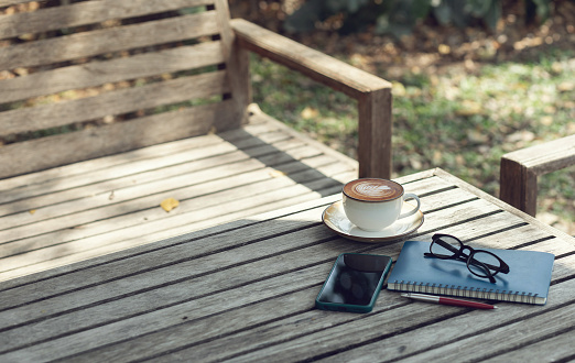 Latte coffee with mobile and note book on old wooden table in outdoor garden