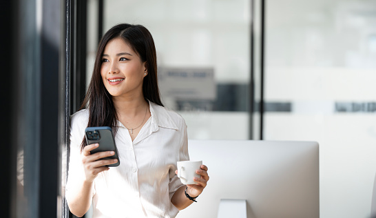 Beautiful asian businesswoman using mobile phone while standing in office, looking up and holding coffee cup.