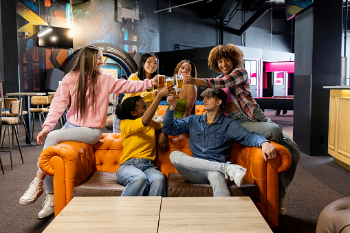 Cheerful friends toasting with beverages at a colorful arcade lounge.