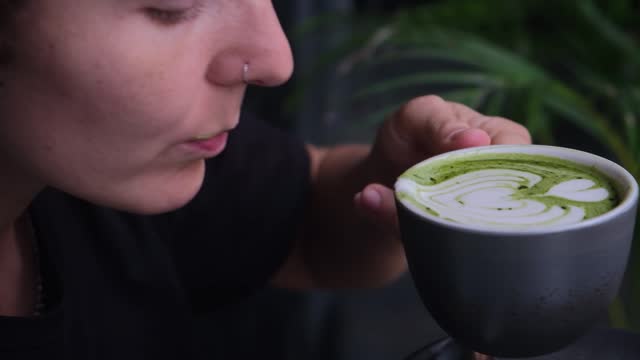 Beautiful woman drinking from mug of trendy matcha green drink with amazing latte art in hipster cafe. Organic green mast is filled with healing powers. Ideal morning concept with green coffee.