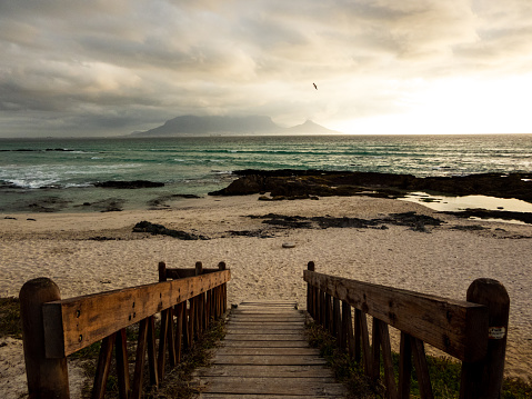 A scenic view of a wooden walkway leading down to a cloudy beach with Table Mountain in the distance