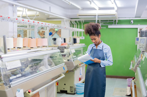 American female designer is a seamstress checking product list waiting to be distributed to regional customers in a weaving industry factory with computerized sewing machines working.