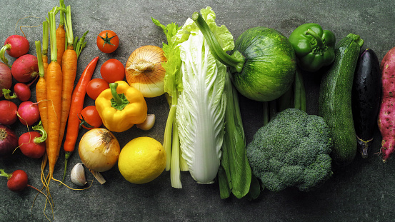 Many market fresh vegetables lined up next to each other in the vibrant colors of a rainbow. Vegetables include; zucchini, broccoli, aubergine, sweet potato radish, red onion, tomato, chili pepper, carrot, bell pepper, onion, garlic, bok choy, cabbage, pumpkin celery, leek, beans and a lemon. Shot from directly above.