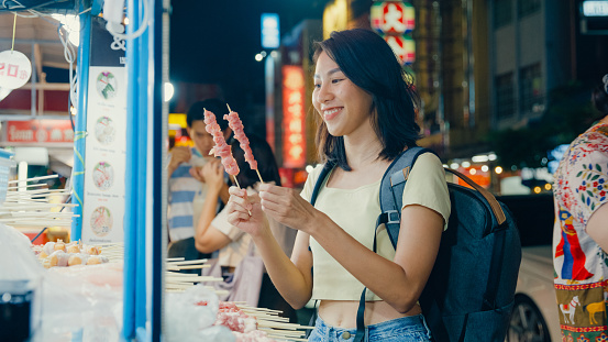 Young Asian woman backpacker enjoy buying street food on the street at night market in Bangkok, Thailand. Holiday vacation trip concept.