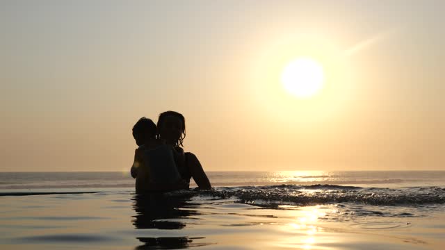 Mother and children swim in warm sea against backdrop of beautiful sunset. Child happily dives into sea surrounded by rays of setting sun. Moments of happiness for whole family by beautiful summer sea