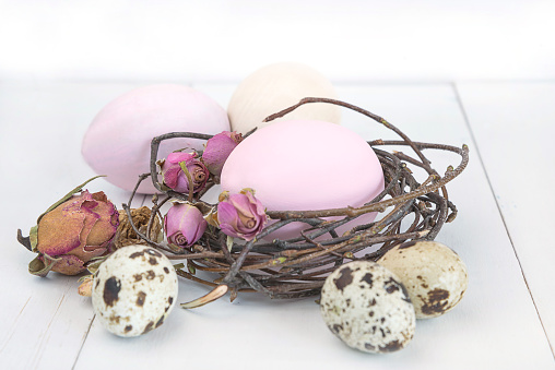 handmade painted eggs and quail eggs in a nest, dry rose flowers on white wooden background