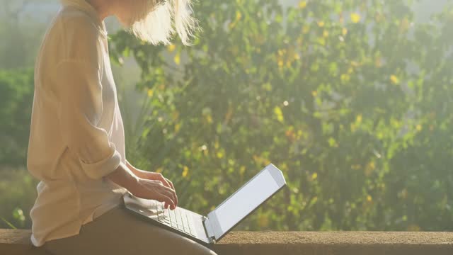 Excited young woman typing on a laptop keyboard in sunshine with beautiful lense flare effect. Female freelancer works on the rooftop outdoor at sunrise or sunset.
