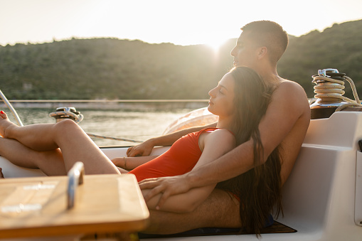 Beautiful young couple relaxing and enjoying together on sailboat cockpit at sunset. They are affectionate, hugging each other, smiling and having a fun. Part of series