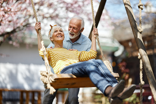 Happy senior man pushing his wife on a swing during spring day in the park. Focus is on woman.