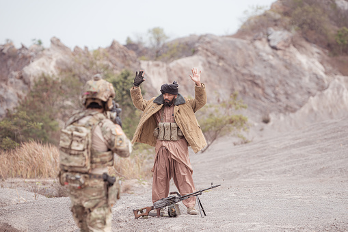 A Taliban soldier stands with a machine gun, Raise hands in surrender to the allied soldiers, In the desert mountain terrain battlefield