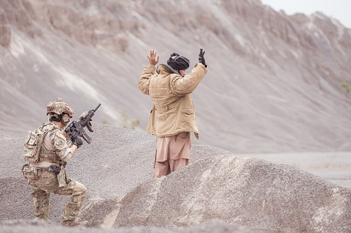 A Taliban soldier stands with a machine gun, Raise hands in surrender to the allied soldiers, In the desert mountain terrain battlefield