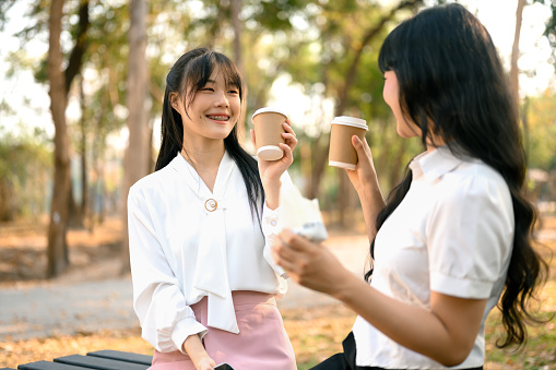 Happy young Asian women drinking coffee from paper cups and chatting with each other in the park.