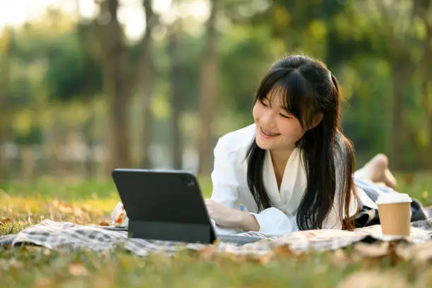 Portrait of pretty young Asian woman lying on picnic blanker and using digital tablet.