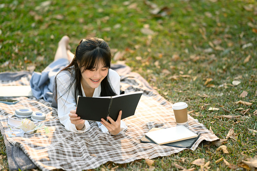 Relaxed young woman lying on picnic blanket and reading boo, enjoying free time at outdoor.
