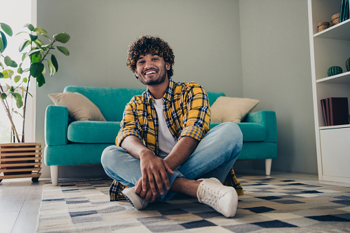 Photo of positive glad nice man freelancer sitting on floor in comfortable flat dorm living room enjoying free time alone indoors.