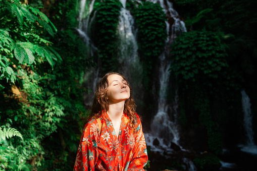 Woman  in red kimono standing near the refreshing waterfall on Bali