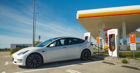 Shell gas station in Croatia, 05.05.2023: CLOSE UP: White Tesla electric car connected to Supercharger post at gas station. Plugged white BEV car efficiently recharging lithium battery in the motor.